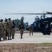 A group of military personnel in full combat gear and parachute equipment are lined up on a tarmac, preparing to board a dark military helicopter, likely a UH-60 Black Hawk. Two individuals in black t-shirts appear to be instructors or jumpmaster personnel guiding the formation toward the aircraft. The scene depicts a pre-jump or air assault operation at a military airfield.