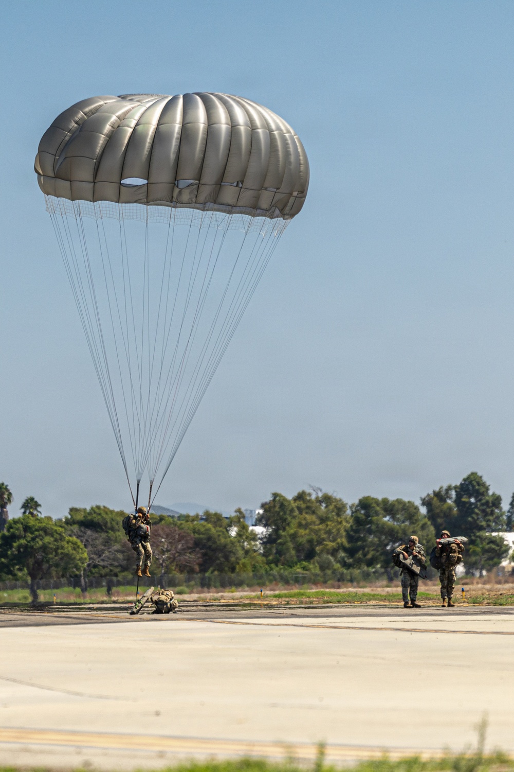 A military paratrooper is landing on a paved airstrip with a fully deployed olive/tan-colored round parachute. Two other soldiers in military gear are standing nearby on the tarmac, observing or assisting. The setting appears to be a military airfield surrounded by trees and vegetation under clear blue skies.