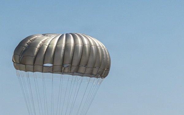A military paratrooper is landing on a paved airstrip with a fully deployed olive/tan-colored round parachute. Two other soldiers in military gear are standing nearby on the tarmac, observing or assisting. The setting appears to be a military airfield surrounded by trees and vegetation under clear blue skies.