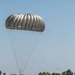 A military paratrooper is landing on a paved airstrip with a fully deployed olive/tan-colored round parachute. Two other soldiers in military gear are standing nearby on the tarmac, observing or assisting. The setting appears to be a military airfield surrounded by trees and vegetation under clear blue skies.