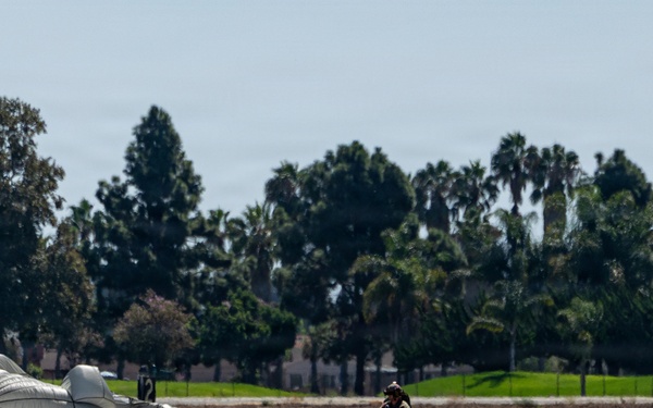 A military paratrooper has just landed on a flat, paved surface, likely an airfield or tarmac. A large white/gray parachute canopy is collapsed and spread across the ground to the left of the jumper. The background features a treeline with palm trees and deciduous trees, suggesting a warm-climate location.