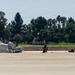 A military paratrooper has just landed on a flat, paved surface, likely an airfield or tarmac. A large white/gray parachute canopy is collapsed and spread across the ground to the left of the jumper. The background features a treeline with palm trees and deciduous trees, suggesting a warm-climate location.