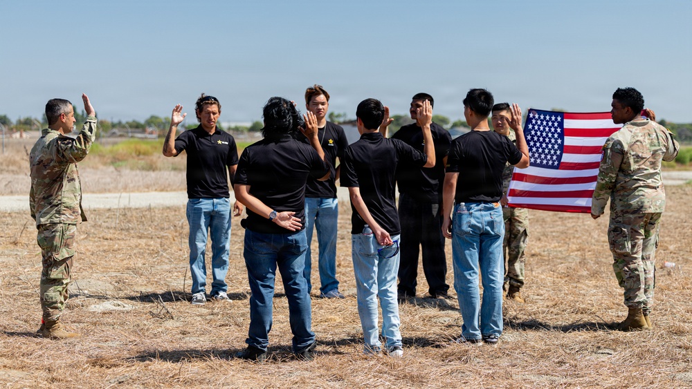 A military oath-taking ceremony is taking place outdoors on a dry grassy field. A uniformed soldier on the left administers an oath to a group of civilians wearing matching black shirts and jeans, while another soldier on the right holds an American flag. This appears to be a military enlistment or naturalization ceremony.