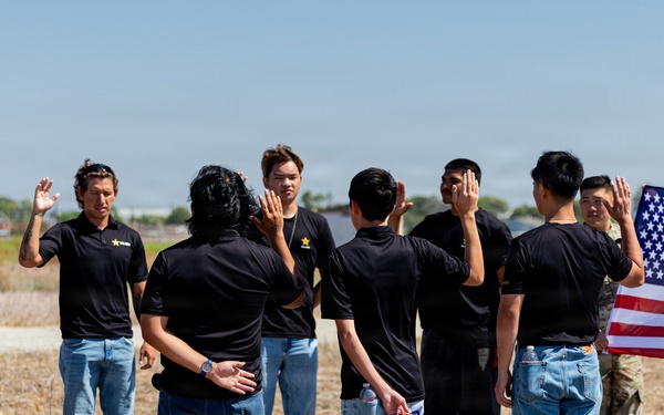 A military oath-taking ceremony is taking place outdoors on a dry grassy field. A uniformed soldier on the left administers an oath to a group of civilians wearing matching black shirts and jeans, while another soldier on the right holds an American flag. This appears to be a military enlistment or naturalization ceremony.