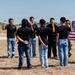 A military oath-taking ceremony is taking place outdoors on a dry grassy field. A uniformed soldier on the left administers an oath to a group of civilians wearing matching black shirts and jeans, while another soldier on the right holds an American flag. This appears to be a military enlistment or naturalization ceremony.