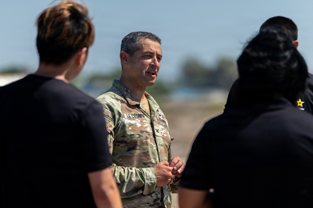 A U.S. Army officer in OCP (Operational Camouflage Pattern) uniform is speaking to a small group of individuals in civilian black t-shirts outdoors. The officer's name tape reads 'LE CEDOP' or similar, and rank insignia is visible on the uniform. The setting appears to be an outdoor military or training environment under bright daylight conditions.