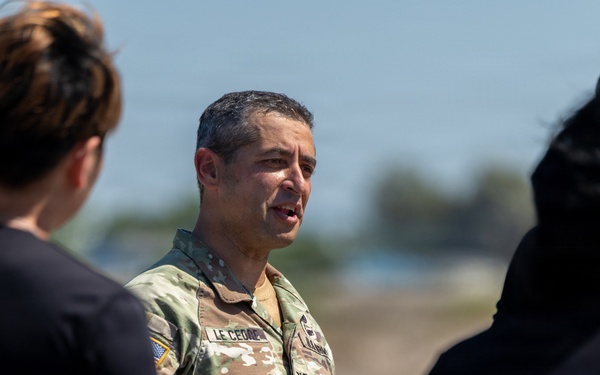 A U.S. Army officer in OCP (Operational Camouflage Pattern) uniform is speaking to a small group of individuals in civilian black t-shirts outdoors. The officer's name tape reads 'LE CEDOP' or similar, and rank insignia is visible on the uniform. The setting appears to be an outdoor military or training environment under bright daylight conditions.