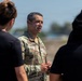 A U.S. Army officer in OCP (Operational Camouflage Pattern) uniform is speaking to a small group of individuals in civilian black t-shirts outdoors. The officer's name tape reads 'LE CEDOP' or similar, and rank insignia is visible on the uniform. The setting appears to be an outdoor military or training environment under bright daylight conditions.