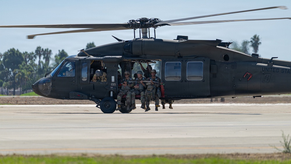 A United States Army UH-60 Black Hawk helicopter is on a tarmac or airfield with soldiers in military camouflage uniforms positioned in and around the open side door. The soldiers appear to be conducting a training exercise or tactical insertion/extraction drill. The setting appears to be a warm-climate military installation, likely in the southwestern United States based on the palm trees visible in the background.