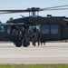 A United States Army UH-60 Black Hawk helicopter is on a tarmac or airfield with soldiers in military camouflage uniforms positioned in and around the open side door. The soldiers appear to be conducting a training exercise or tactical insertion/extraction drill. The setting appears to be a warm-climate military installation, likely in the southwestern United States based on the palm trees visible in the background.