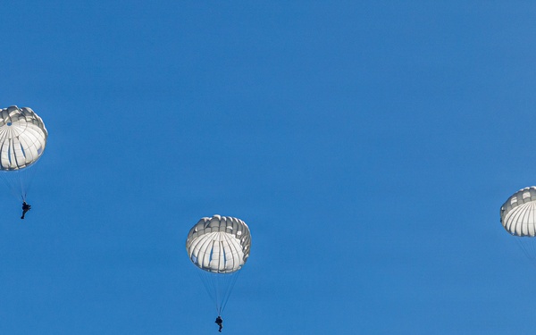 Three paratroopers are descending through a clear blue sky using round, white military-style parachutes. The parachutes appear to be standard round canopy designs consistent with military static-line jumps. The image is taken from below, silhouetting the jumpers against a cloudless sky.