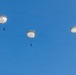 Three paratroopers are descending through a clear blue sky using round, white military-style parachutes. The parachutes appear to be standard round canopy designs consistent with military static-line jumps. The image is taken from below, silhouetting the jumpers against a cloudless sky.