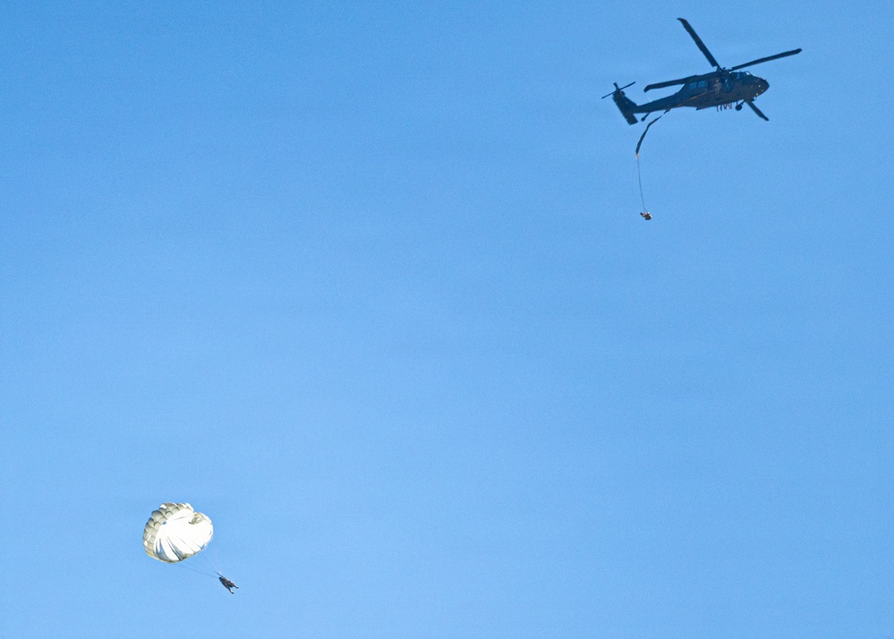 A military helicopter, appearing to be a UH-60 Black Hawk, is flying in a clear blue sky with a person or object suspended below it on a line. Separately, a parachutist with a deployed round parachute is descending in the lower left portion of the frame. The scene appears to depict a military training or operational exercise involving both helicopter operations and parachuting.