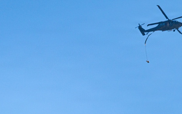 A military helicopter, appearing to be a UH-60 Black Hawk, is flying in a clear blue sky with a person or object suspended below it on a line. Separately, a parachutist with a deployed round parachute is descending in the lower left portion of the frame. The scene appears to depict a military training or operational exercise involving both helicopter operations and parachuting.
