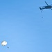 A military helicopter, appearing to be a UH-60 Black Hawk, is flying in a clear blue sky with a person or object suspended below it on a line. Separately, a parachutist with a deployed round parachute is descending in the lower left portion of the frame. The scene appears to depict a military training or operational exercise involving both helicopter operations and parachuting.