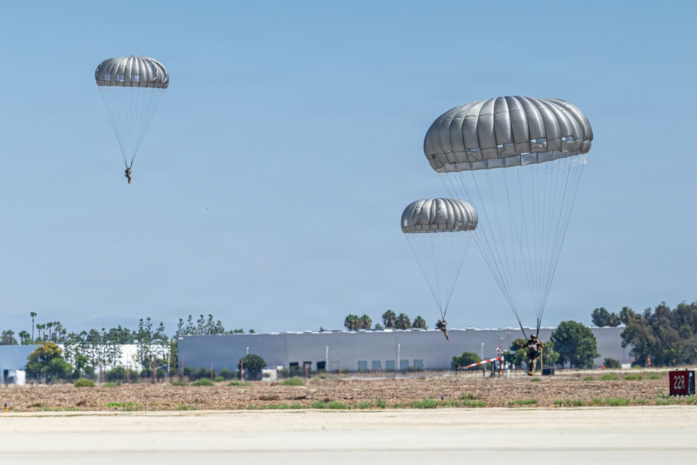 Three military paratroopers are descending with round, gray/silver parachutes over what appears to be an airfield or military installation. One paratrooper is near ground level and appears to be landing, while the other two are at higher altitudes. The setting appears to be a sunny day at a facility with large warehouse-style buildings and trees visible in the background.