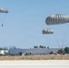 Three military paratroopers are descending with round, gray/silver parachutes over what appears to be an airfield or military installation. One paratrooper is near ground level and appears to be landing, while the other two are at higher altitudes. The setting appears to be a sunny day at a facility with large warehouse-style buildings and trees visible in the background.