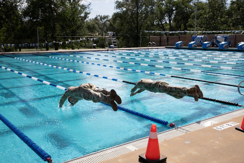 Two individuals wearing military camouflage uniforms and boots are captured mid-air as they dive into an outdoor competitive swimming pool. The image appears to document a military water training exercise, as both subjects are fully clothed in combat attire rather than swimwear. The pool is a standard multi-lane competition facility with blue lane dividers, visible depth markings, and surrounding infrastructure.
