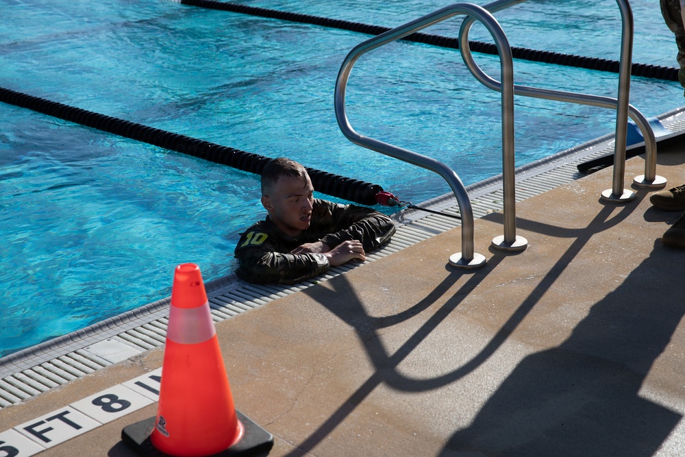 A person wearing camouflage military-style clothing with the number '10' visible is in a swimming pool, holding onto the pool edge near a metal ladder/handrail. The pool appears to be an outdoor facility marked with depth indicators showing '8 FT'. An orange traffic cone is placed on the pool deck nearby, suggesting an organized training or assessment event.