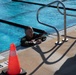 A person wearing camouflage military-style clothing with the number '10' visible is in a swimming pool, holding onto the pool edge near a metal ladder/handrail. The pool appears to be an outdoor facility marked with depth indicators showing '8 FT'. An orange traffic cone is placed on the pool deck nearby, suggesting an organized training or assessment event.