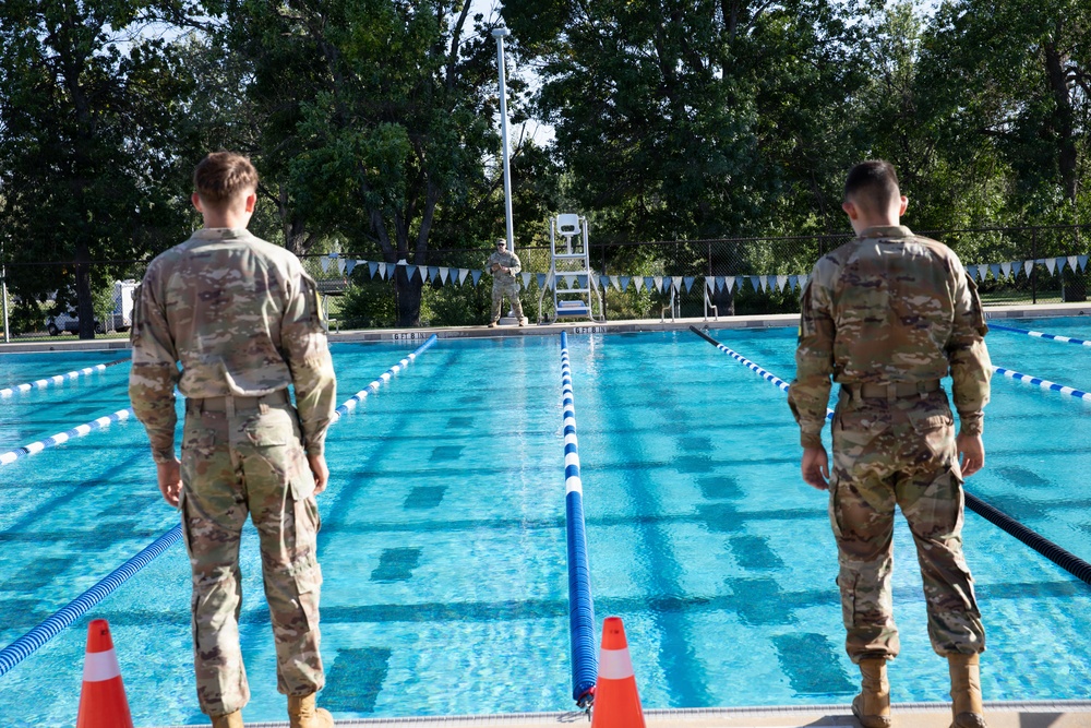 Two military personnel in camouflage uniforms stand at the edge of an outdoor swimming pool with lane dividers. A third uniformed individual is visible at the far end of the pool near a lifeguard chair. The setting appears to be a military installation pool facility, possibly during a swim assessment or training event.