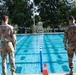 Two military personnel in camouflage uniforms stand at the edge of an outdoor swimming pool with lane dividers. A third uniformed individual is visible at the far end of the pool near a lifeguard chair. The setting appears to be a military installation pool facility, possibly during a swim assessment or training event.