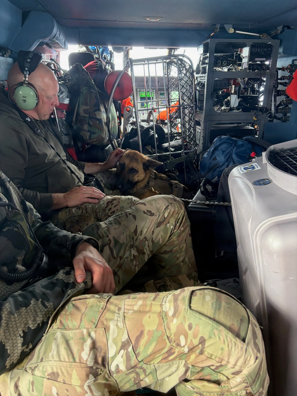 Military or law enforcement personnel in camouflage uniforms are seated inside what appears to be a helicopter or large military aircraft. A Belgian Malinois or German Shepherd dog wearing a tactical harness is resting on one of the personnel's lap. The interior shows aviation equipment, a metal cage/crate, storage containers, and communications gear consistent with a search and rescue or tactical operation.