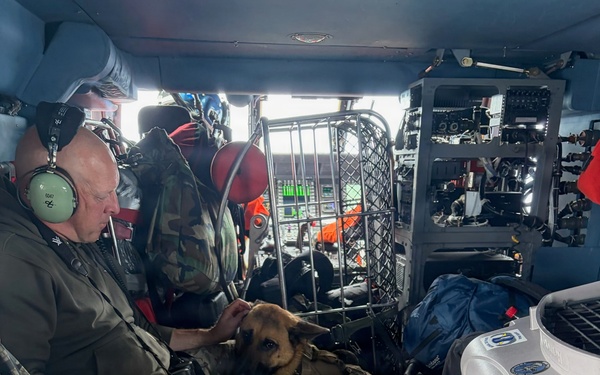 Military or law enforcement personnel in camouflage uniforms are seated inside what appears to be a helicopter or large military aircraft. A Belgian Malinois or German Shepherd dog wearing a tactical harness is resting on one of the personnel's lap. The interior shows aviation equipment, a metal cage/crate, storage containers, and communications gear consistent with a search and rescue or tactical operation.