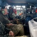 Military or law enforcement personnel in camouflage uniforms are seated inside what appears to be a helicopter or large military aircraft. A Belgian Malinois or German Shepherd dog wearing a tactical harness is resting on one of the personnel's lap. The interior shows aviation equipment, a metal cage/crate, storage containers, and communications gear consistent with a search and rescue or tactical operation.