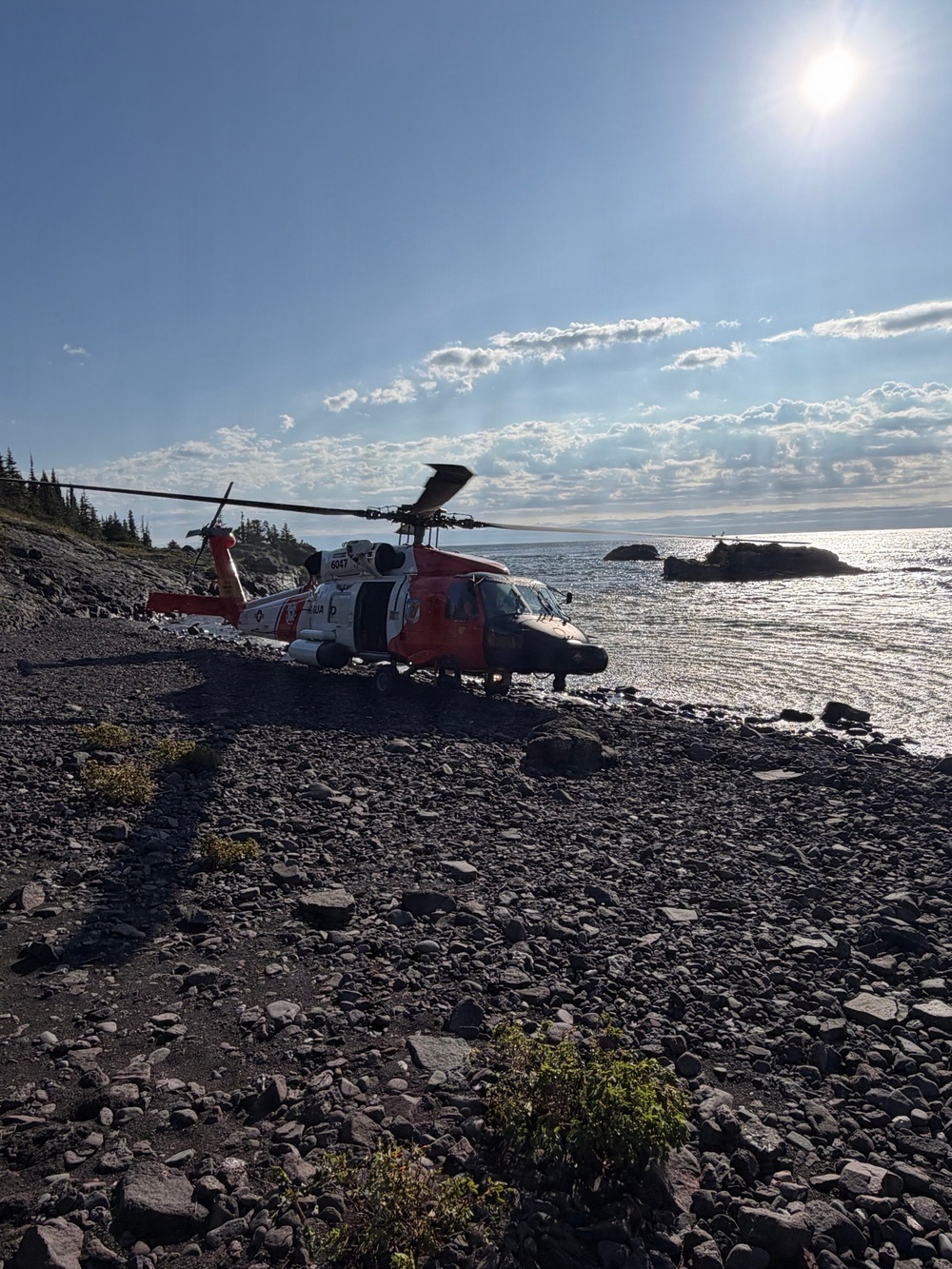 A U.S. Coast Guard MH-60 Jayhawk helicopter (tail number 6047) is landed on a rocky coastal shoreline. The aircraft displays the distinctive orange and white Coast Guard color scheme with the racing stripe insignia. The setting appears to be a remote, rugged coastline with calm water and scattered rocks visible in the background.