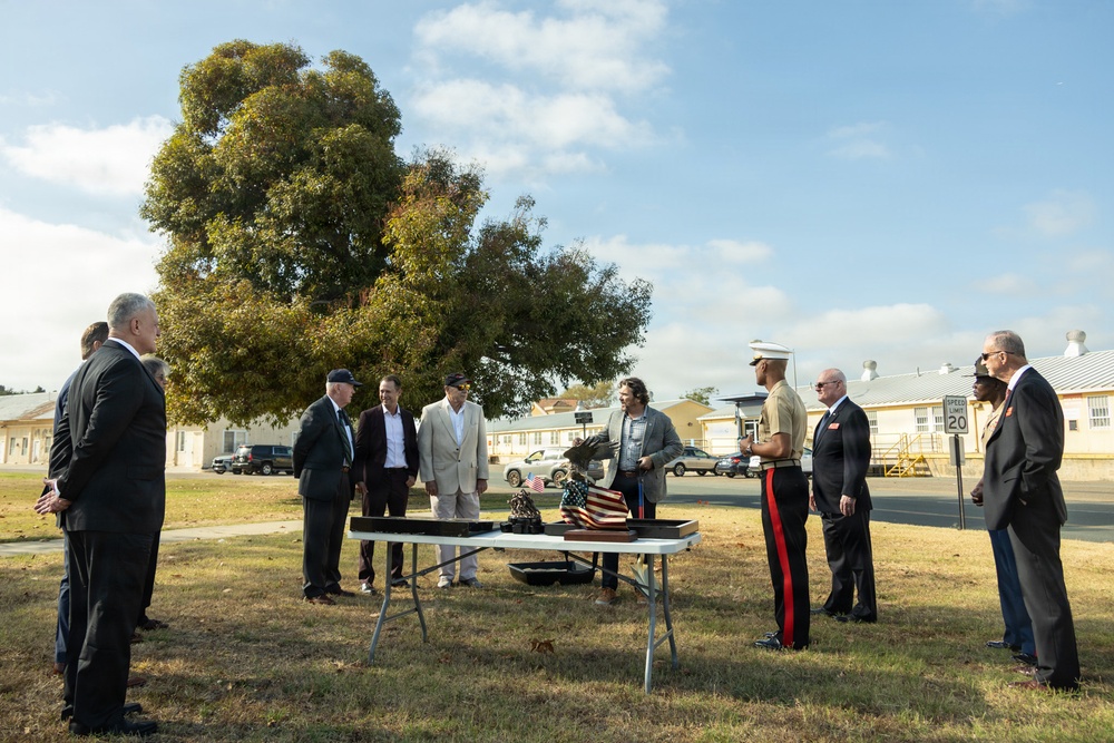 A small outdoor ceremony is taking place on a military installation, with approximately ten individuals gathered around a folding table displaying what appears to be a folded American flag and a small statue or trophy. One participant is wearing a United States Marine Corps dress uniform with red stripe trousers, while others are dressed in civilian business attire. The setting includes military-style barracks buildings in the background and a speed limit sign reading 20 mph.