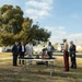 A small outdoor ceremony is taking place on a military installation, with approximately ten individuals gathered around a folding table displaying what appears to be a folded American flag and a small statue or trophy. One participant is wearing a United States Marine Corps dress uniform with red stripe trousers, while others are dressed in civilian business attire. The setting includes military-style barracks buildings in the background and a speed limit sign reading 20 mph.