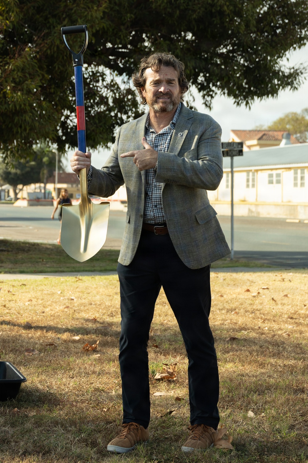 A man in business casual attire stands outdoors on a grass area holding a ceremonial gold shovel with a blue and red handle, pointing at it with his free hand and smiling. The setting appears to be a groundbreaking ceremony, likely at a public or institutional location. A street sign, buildings, and trees are visible in the background.