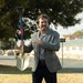A man in business casual attire stands outdoors on a grass area holding a ceremonial gold shovel with a blue and red handle, pointing at it with his free hand and smiling. The setting appears to be a groundbreaking ceremony, likely at a public or institutional location. A street sign, buildings, and trees are visible in the background.