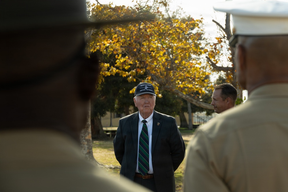 An elderly man wearing a dark blazer, striped tie, and a military-style cap stands outdoors addressing what appears to be a group of uniformed military personnel. The scene is framed by two blurred figures in the foreground wearing tan/khaki uniforms, with one wearing a white cover (cap). The setting appears to be a military installation or park with autumn-colored trees in the background.