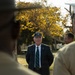 An elderly man wearing a dark blazer, striped tie, and a military-style cap stands outdoors addressing what appears to be a group of uniformed military personnel. The scene is framed by two blurred figures in the foreground wearing tan/khaki uniforms, with one wearing a white cover (cap). The setting appears to be a military installation or park with autumn-colored trees in the background.