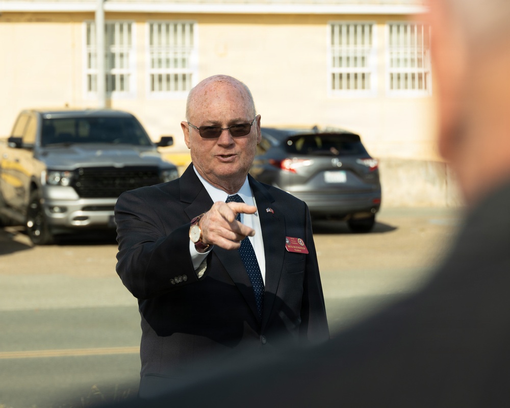 An older bald man wearing a dark suit, tie, and sunglasses is pointing toward the camera or a nearby subject. He is wearing a red name badge and an American flag pin on his lapel, suggesting an official or formal role. The setting appears to be an outdoor parking lot near a light-colored institutional building.
