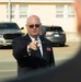 An older bald man wearing a dark suit, tie, and sunglasses is pointing toward the camera or a nearby subject. He is wearing a red name badge and an American flag pin on his lapel, suggesting an official or formal role. The setting appears to be an outdoor parking lot near a light-colored institutional building.