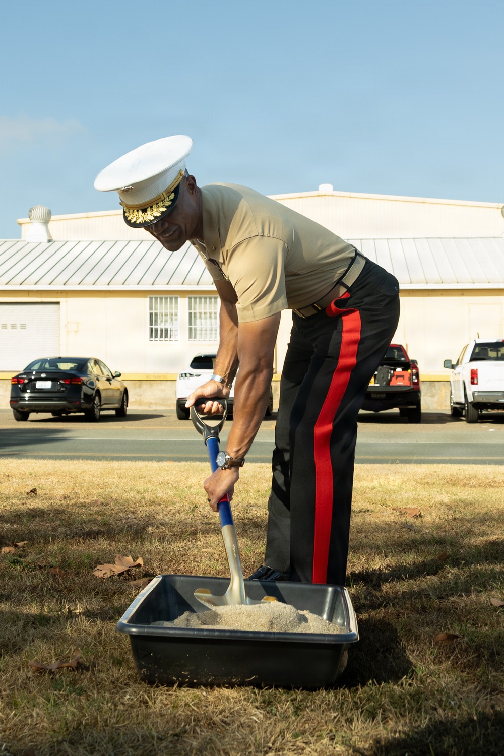 A senior U.S. Marine Corps officer in service uniform performs a ceremonial groundbreaking, shoveling sand from a black tray with a ceremonial shovel. The officer wears the distinctive Marine Corps service uniform with black trousers featuring a red 'blood stripe,' a tan service shirt, and a white peaked cap with gold oak leaf embellishments indicating general officer rank. The setting appears to be a military installation with a building and parked vehicles visible in the background.
