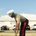 A senior U.S. Marine Corps officer in service uniform performs a ceremonial groundbreaking, shoveling sand from a black tray with a ceremonial shovel. The officer wears the distinctive Marine Corps service uniform with black trousers featuring a red 'blood stripe,' a tan service shirt, and a white peaked cap with gold oak leaf embellishments indicating general officer rank. The setting appears to be a military installation with a building and parked vehicles visible in the background.