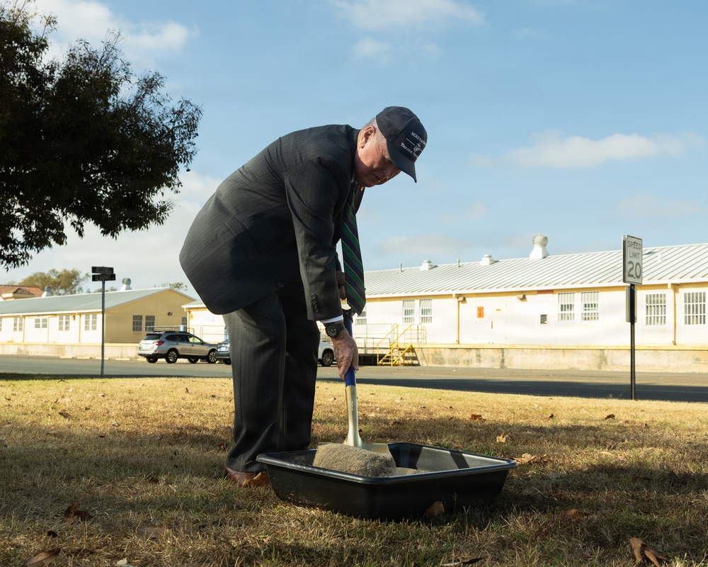 An elderly man in a dark suit and striped tie, wearing a dark baseball cap, is shoveling dirt or sand into a black plastic tray outdoors. The setting appears to be a groundbreaking ceremony on what looks like a military installation or institutional campus, based on the yellow buildings and speed limit sign visible in the background. The scene is photographed in daylight with clear skies.
