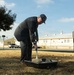 An elderly man in a dark suit and striped tie, wearing a dark baseball cap, is shoveling dirt or sand into a black plastic tray outdoors. The setting appears to be a groundbreaking ceremony on what looks like a military installation or institutional campus, based on the yellow buildings and speed limit sign visible in the background. The scene is photographed in daylight with clear skies.