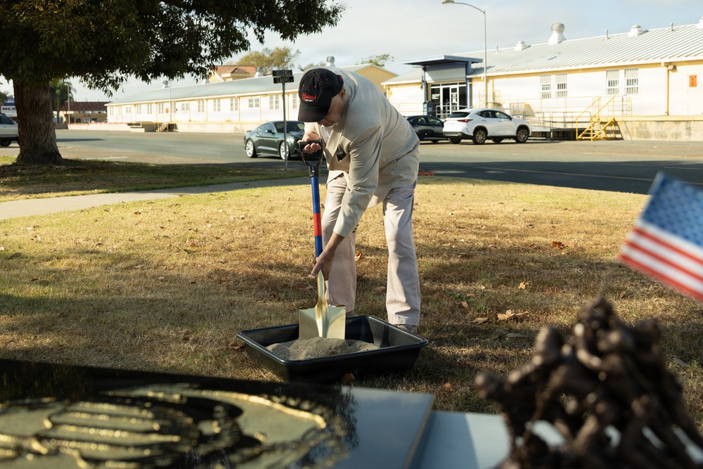 A person wearing a baseball cap and light-colored clothing is using a ceremonial gold-tipped shovel to scoop sand or dirt from a black tray, consistent with a groundbreaking ceremony. An American flag is visible in the foreground right, along with what appears to be ceremonial items. The setting appears to be a military installation or government facility based on the institutional buildings and infrastructure visible in the background.