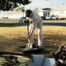 A person wearing a baseball cap and light-colored clothing is using a ceremonial gold-tipped shovel to scoop sand or dirt from a black tray, consistent with a groundbreaking ceremony. An American flag is visible in the foreground right, along with what appears to be ceremonial items. The setting appears to be a military installation or government facility based on the institutional buildings and infrastructure visible in the background.