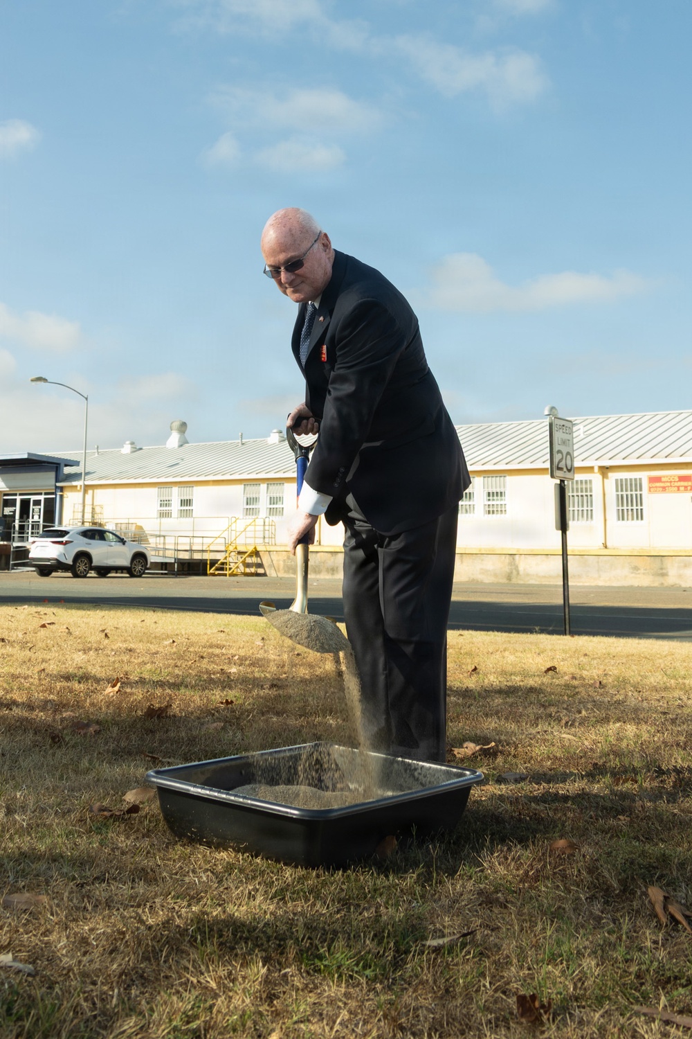 A man in a dark business suit is performing a ceremonial groundbreaking, shoveling dirt into a black plastic tray on a dry grass lawn. The setting appears to be a military or government installation, indicated by the institutional buildings, speed limit sign, and overall environment. The event is taking place outdoors on a clear, partly cloudy day.