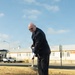 A man in a dark business suit is performing a ceremonial groundbreaking, shoveling dirt into a black plastic tray on a dry grass lawn. The setting appears to be a military or government installation, indicated by the institutional buildings, speed limit sign, and overall environment. The event is taking place outdoors on a clear, partly cloudy day.
