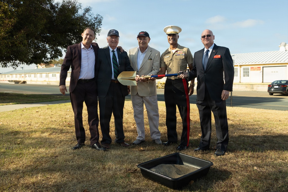 Five men pose together at what appears to be a groundbreaking ceremony, holding a ceremonial gold shovel. One individual is wearing a United States Marine Corps dress uniform with a white cover and service ribbons, while the others are dressed in civilian business attire. A black tray filled with sand or soil is placed in the foreground, consistent with a formal groundbreaking event.