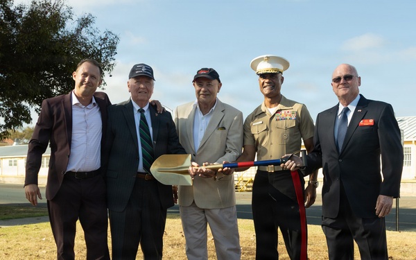 Five men pose together at what appears to be a groundbreaking ceremony, holding a ceremonial gold shovel. One individual is wearing a United States Marine Corps dress uniform with a white cover and service ribbons, while the others are dressed in civilian business attire. A black tray filled with sand or soil is placed in the foreground, consistent with a formal groundbreaking event.