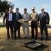 Five men pose together at what appears to be a groundbreaking ceremony, holding a ceremonial gold shovel. One individual is wearing a United States Marine Corps dress uniform with a white cover and service ribbons, while the others are dressed in civilian business attire. A black tray filled with sand or soil is placed in the foreground, consistent with a formal groundbreaking event.
