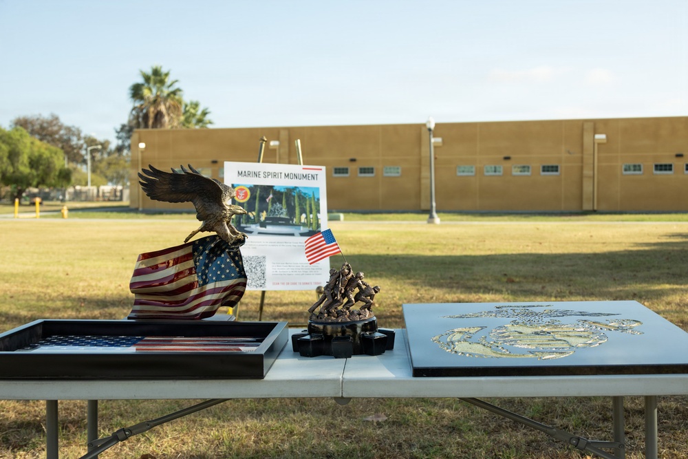 An outdoor display table set up for a Marine Spirit Monument event, featuring patriotic and military memorabilia. The table holds bronze sculptures, American flag items, and a USMC Eagle Globe and Anchor emblem plaque. A sign in the background reads 'Marine Spirit Monument' with a QR code, set against a military-style building.